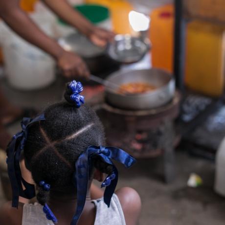 A young girl watches as a woman prepares food in a displacement camp in Haiti